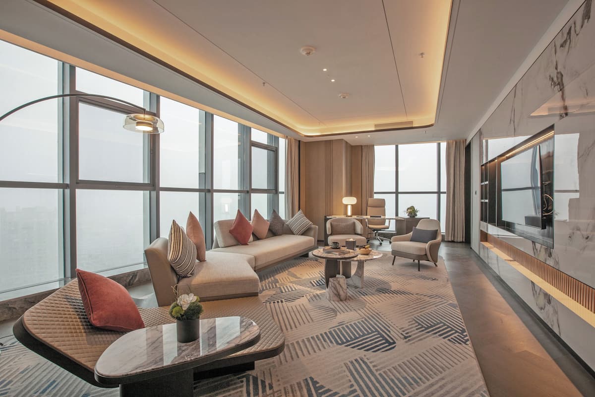 A modern hotel room with a large window, a sofa, a coffee table, and a TV.
