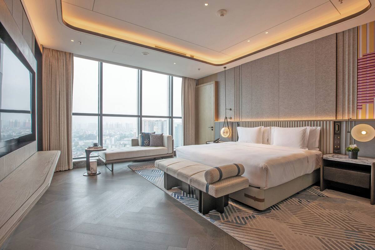 A modern hotel room with a large bed, a bench, a bedside table, and a view of the city through large windows.