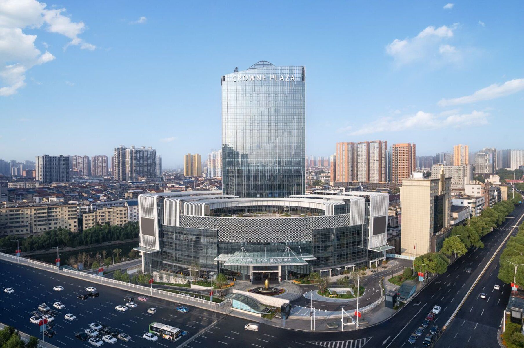 A modern building with a glass facade and a circular lower section, surrounded by a parking area and roads.
