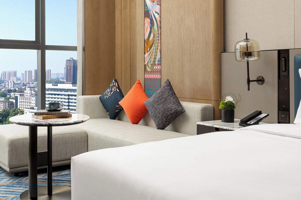A hotel room with a large bed, a sofa, a small table with a book, a potted plant, and a city view.