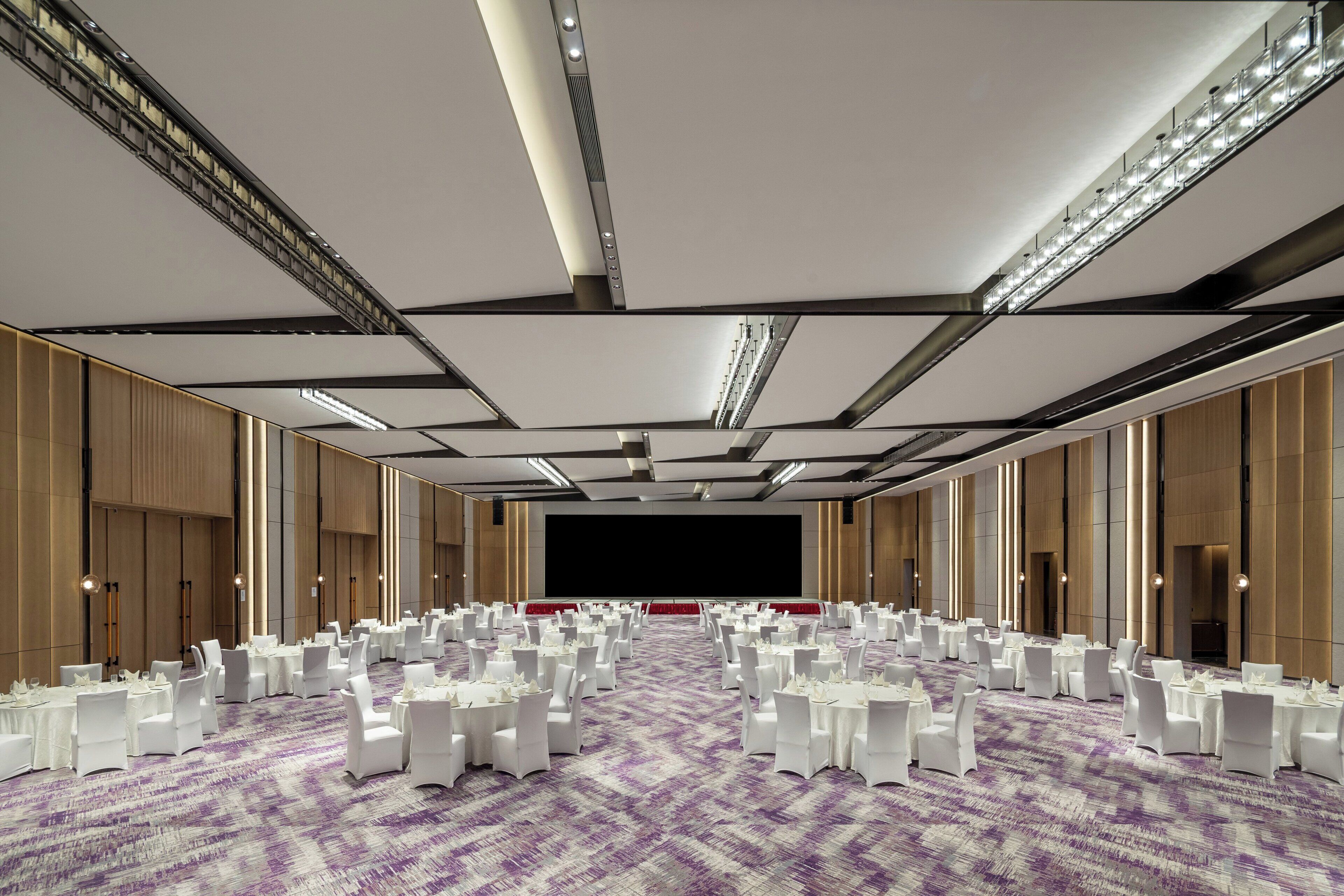 A large conference room with rows of round tables and chairs, a stage, and a high ceiling with lighting fixtures.