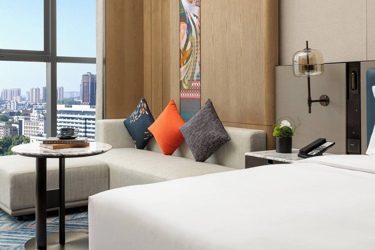 A hotel room with a large bed, a sofa, a small table with a book, a potted plant, and a city view.