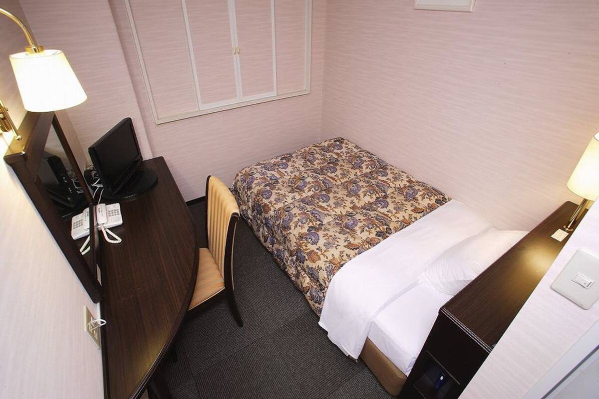 A small, neatly arranged hotel room with a bed, a desk, a chair, and a lamp.