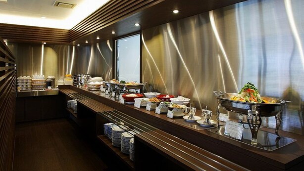 A buffet setup with various food items and serving utensils, a wooden counter, and a metallic backdrop. - Hamamatsu Hotel (Hamamatsu)