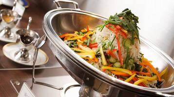 A stainless steel buffet bowl filled with a colorful vegetable stir-fry and rice, accompanied by small bowls of sauces.