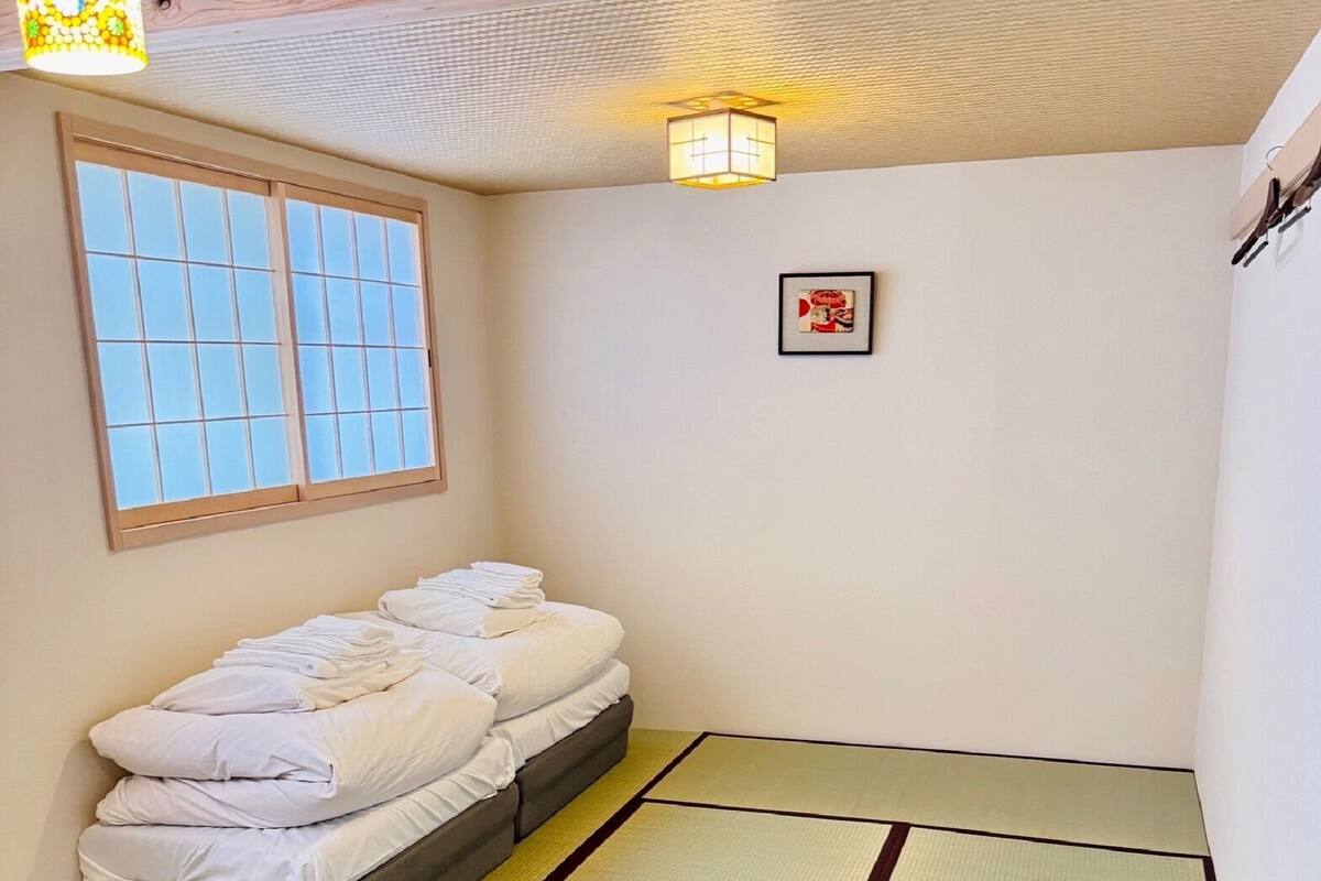 A small room with a bed, a window, a wall-mounted light, and a framed picture.