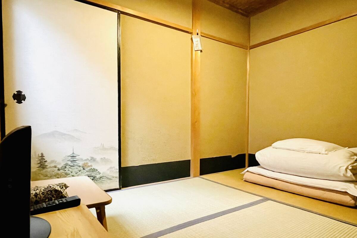A traditional Japanese room with tatami flooring, a futon bed, a low wooden desk, and a sliding door with a landscape painting.
