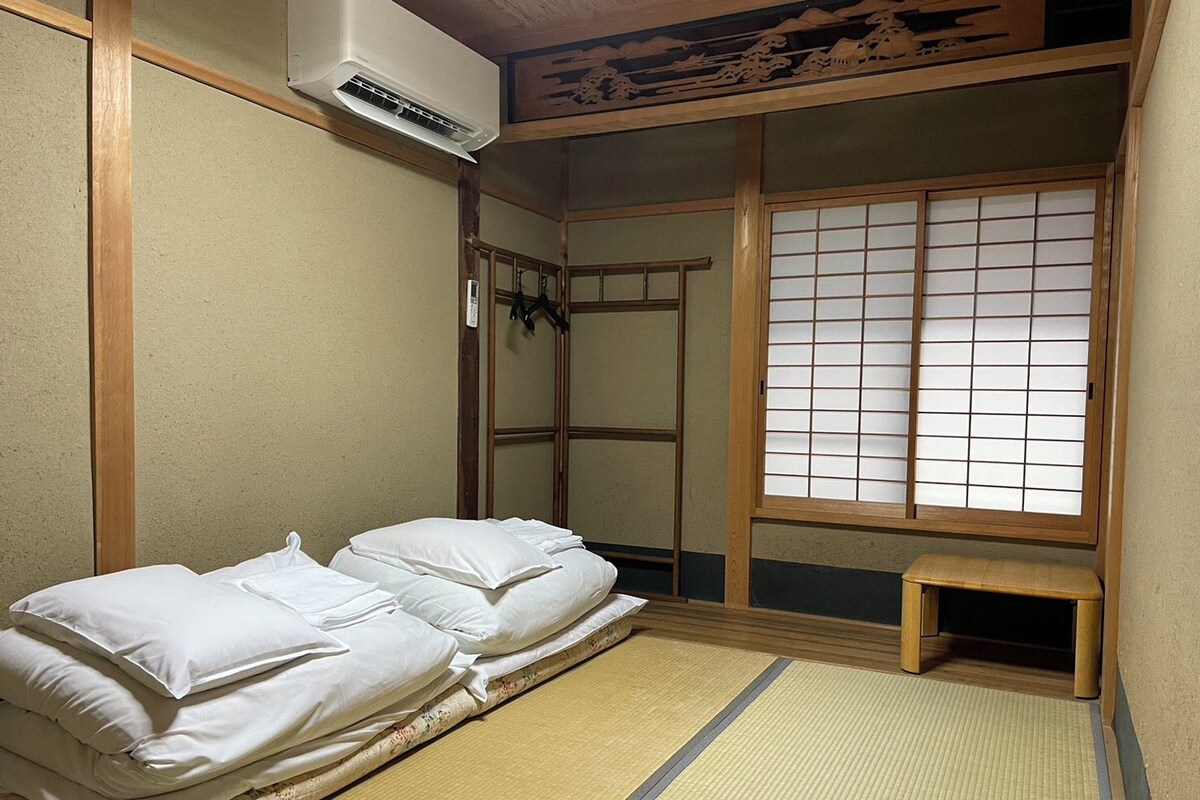 Una habitación tradicional japonesa con suelo de tatami, futón, pantalla shoji y un pequeño banco de madera.