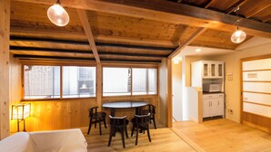 A room with a wooden ceiling, a dining table with chairs, a bed, a window with blinds, and a kitchen area with cabinets.