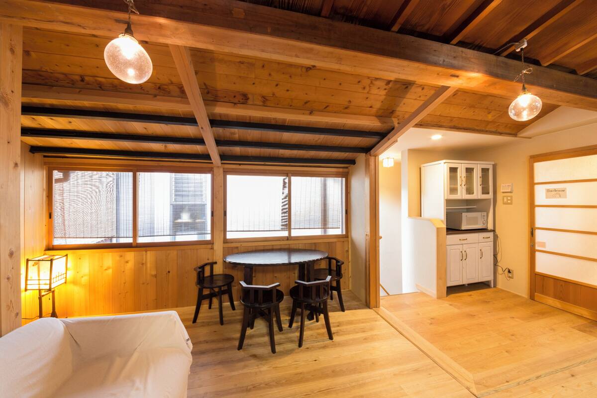 A room with a wooden ceiling, a dining table with chairs, a bed, a window with blinds, and a kitchen area with cabinets.