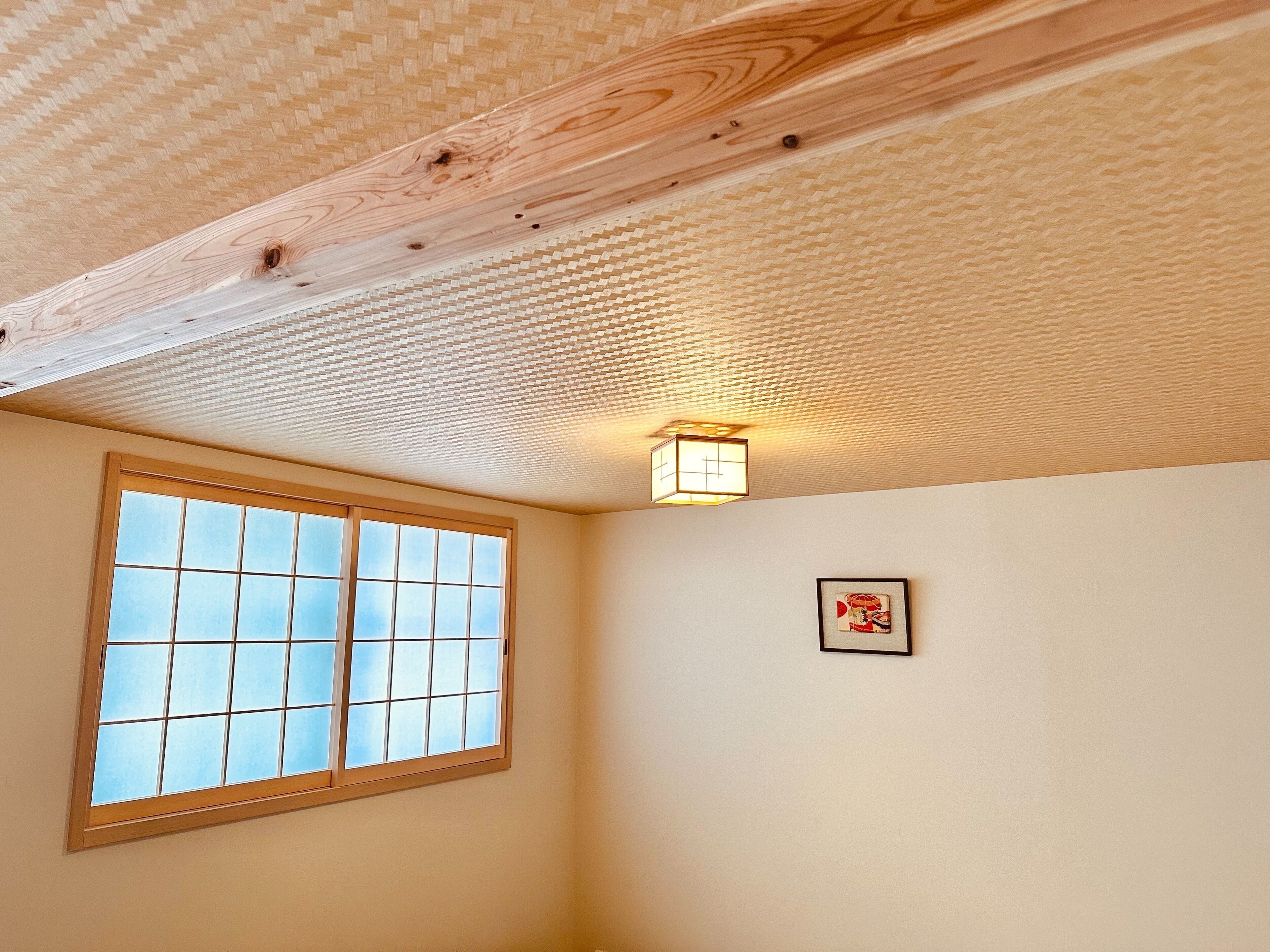 A room with a wooden ceiling, a window with a grid pattern, and a wall-mounted light fixture.