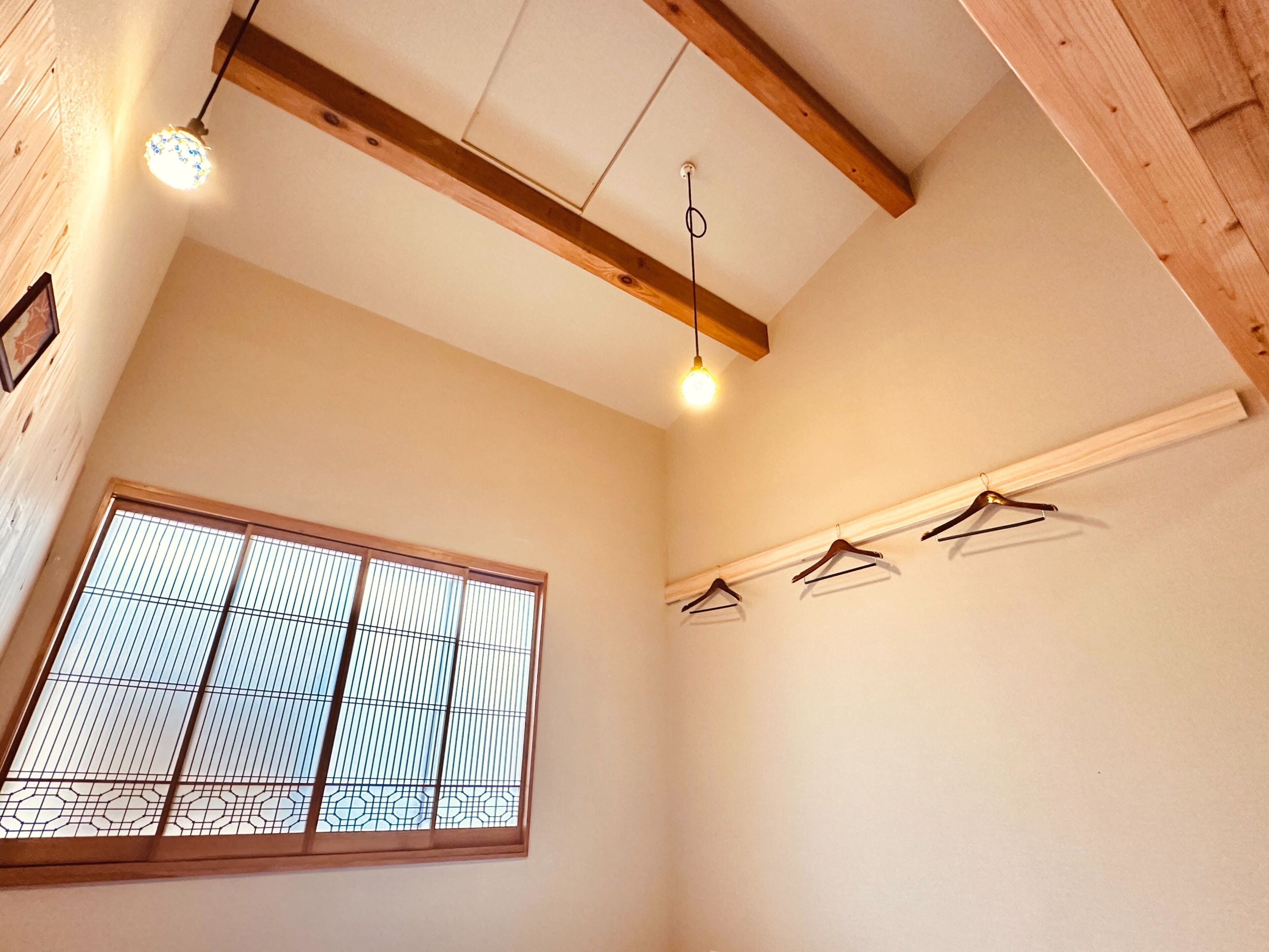 Een kamer met een houten plafond, een raam met een rasterpatroon en drie kledinghaken aan de muur.