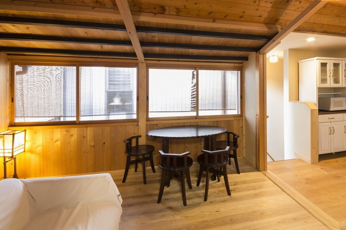 Un interior de madera con una mesa y sillas de comedor, una zona de cocina y un ventanal con persianas.