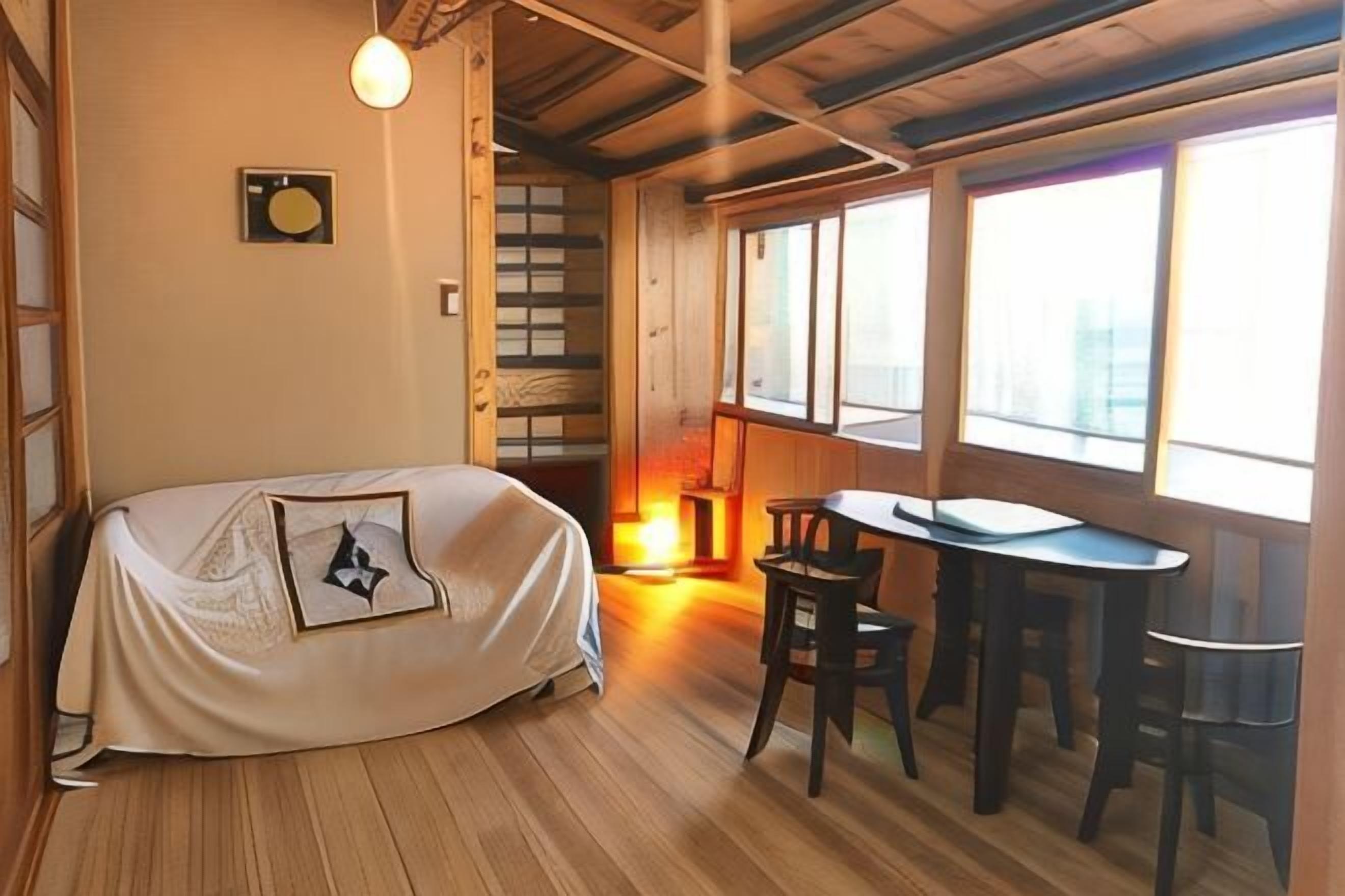 A room with a bed, a table, and chairs, wooden floors, and large windows.