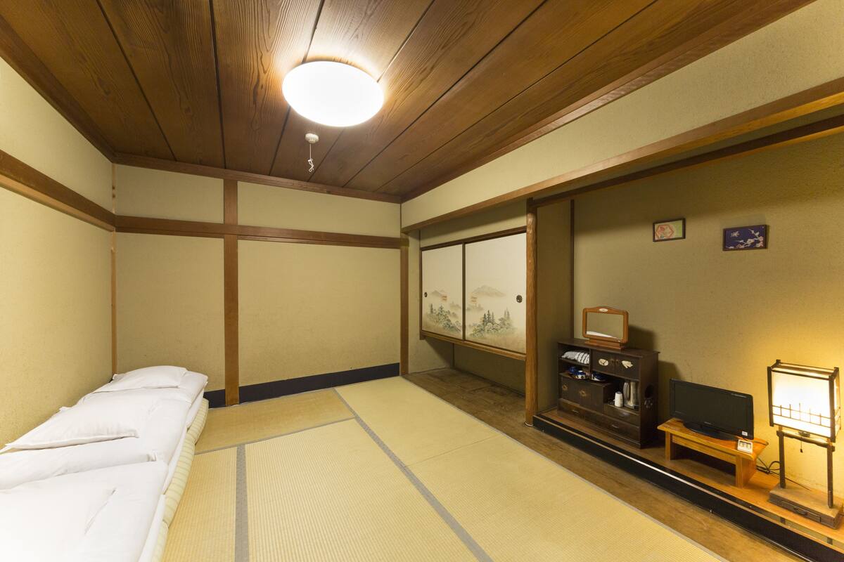 A traditional Japanese room with tatami flooring, a futon bed, a television, and a sliding door with a view of a mountain landscape.