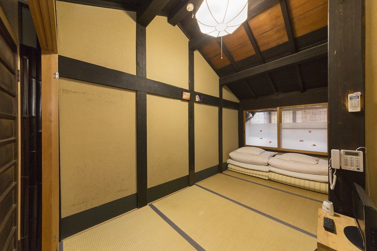 A traditional tatami mat room with a wooden ceiling, a bed with pillows, a telephone, and a small table with a cup.