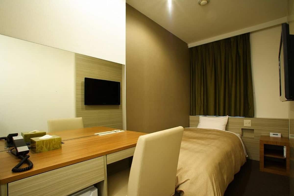 Een hotelkamer met een houten bureau, een stoel, een bed met een beige sprei, een aan de muur bevestigde televisie en een klein nachtkastje.