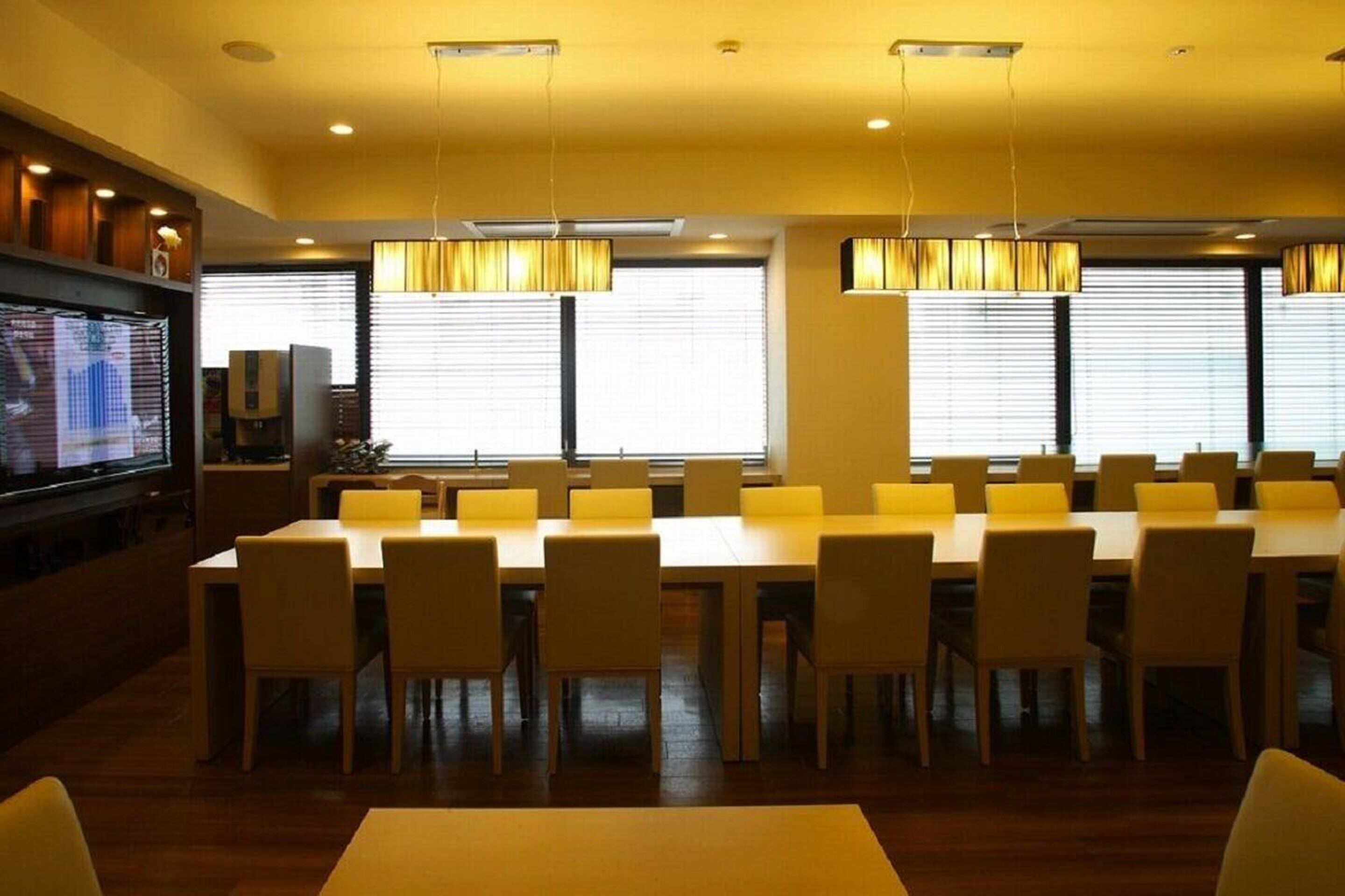 Un salón de conferencias con una larga mesa y sillas, ventanas amplias con persianas y lámparas colgantes.