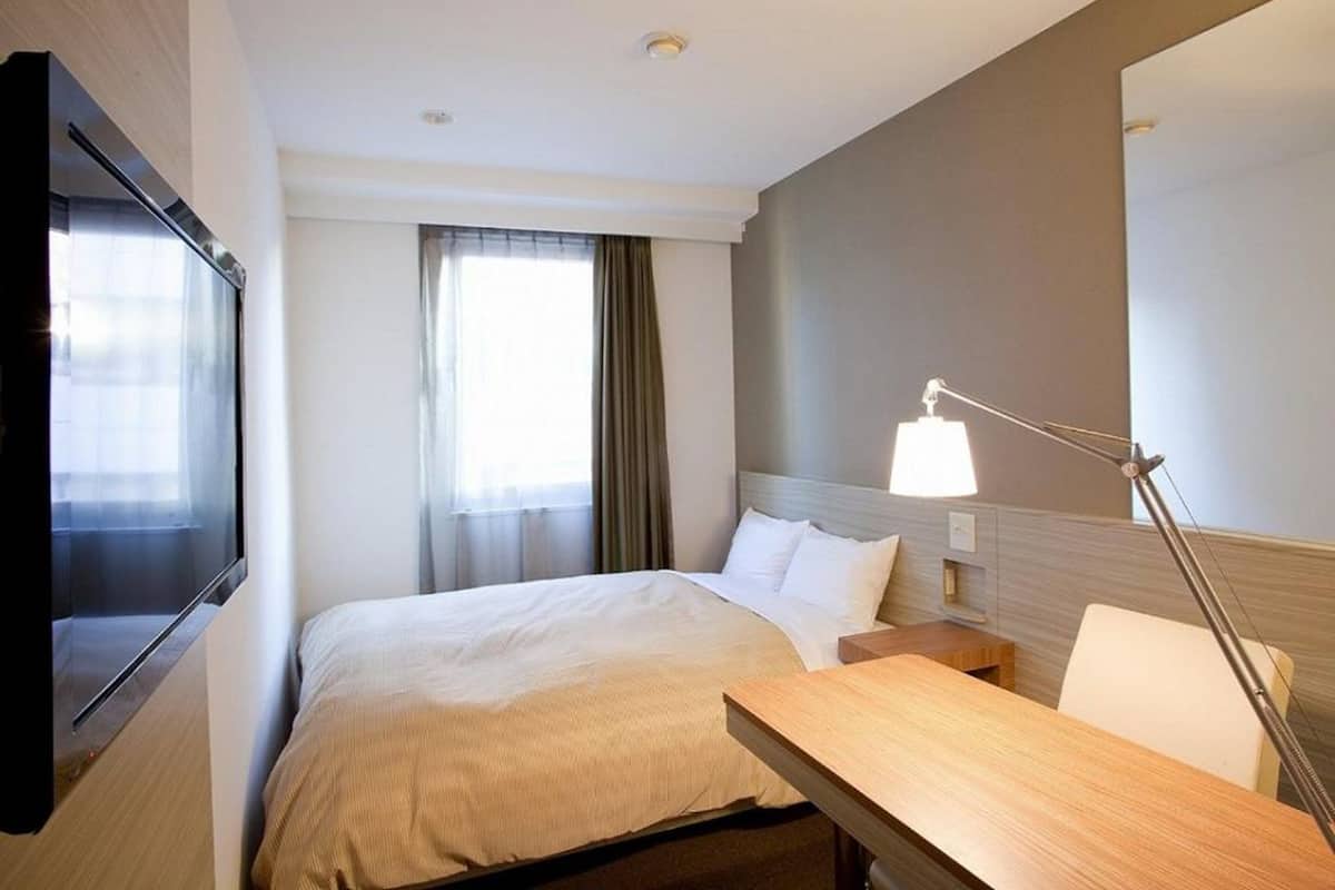 Een hotelkamer met een bed, een bureau, een lamp, een televisie en een raam met gordijnen.