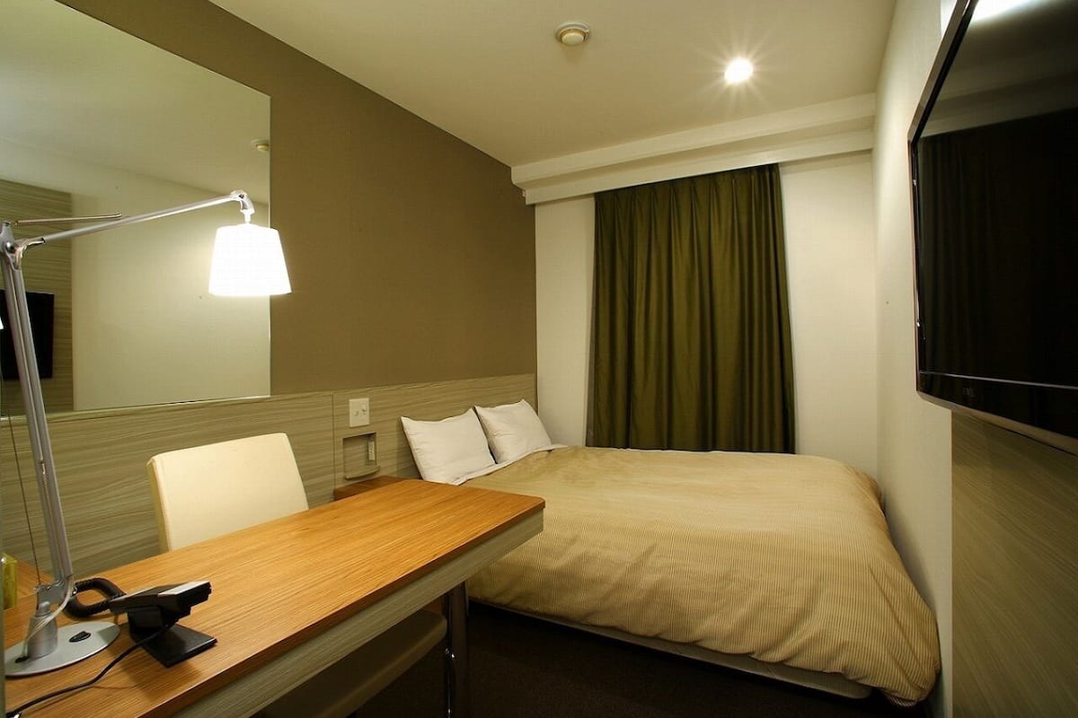 Een hotelkamer met een bed, een bureau met lamp, een stoel, een televisie en een raam met gordijnen.