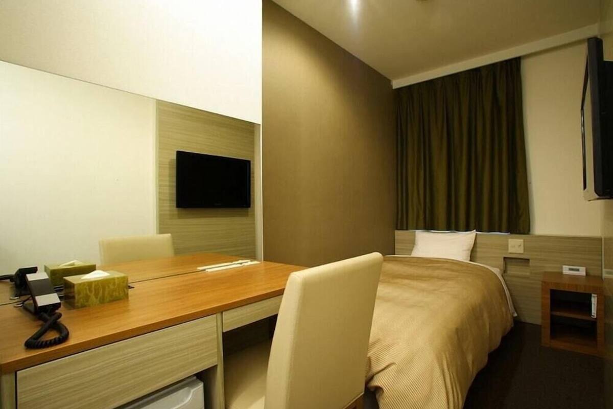 Ein Hotelzimmer mit einem Bett, einem Schreibtisch mit Telefon, einem Sessel, einem an der Wand befestigten Fernseher und einem kleinen Nachttisch.