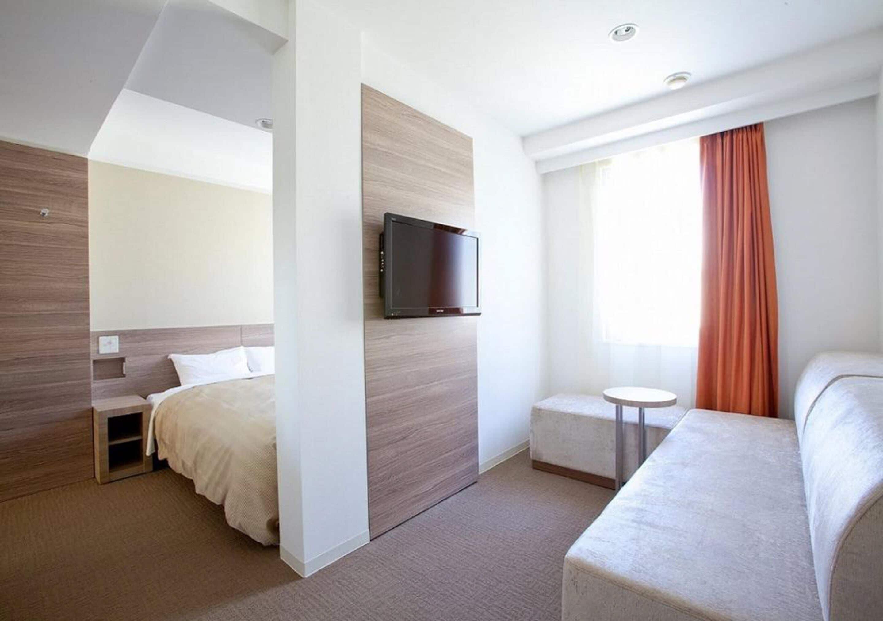 A modern hotel room with a bed, a TV mounted on a wooden panel, a small round table, and a sofa.