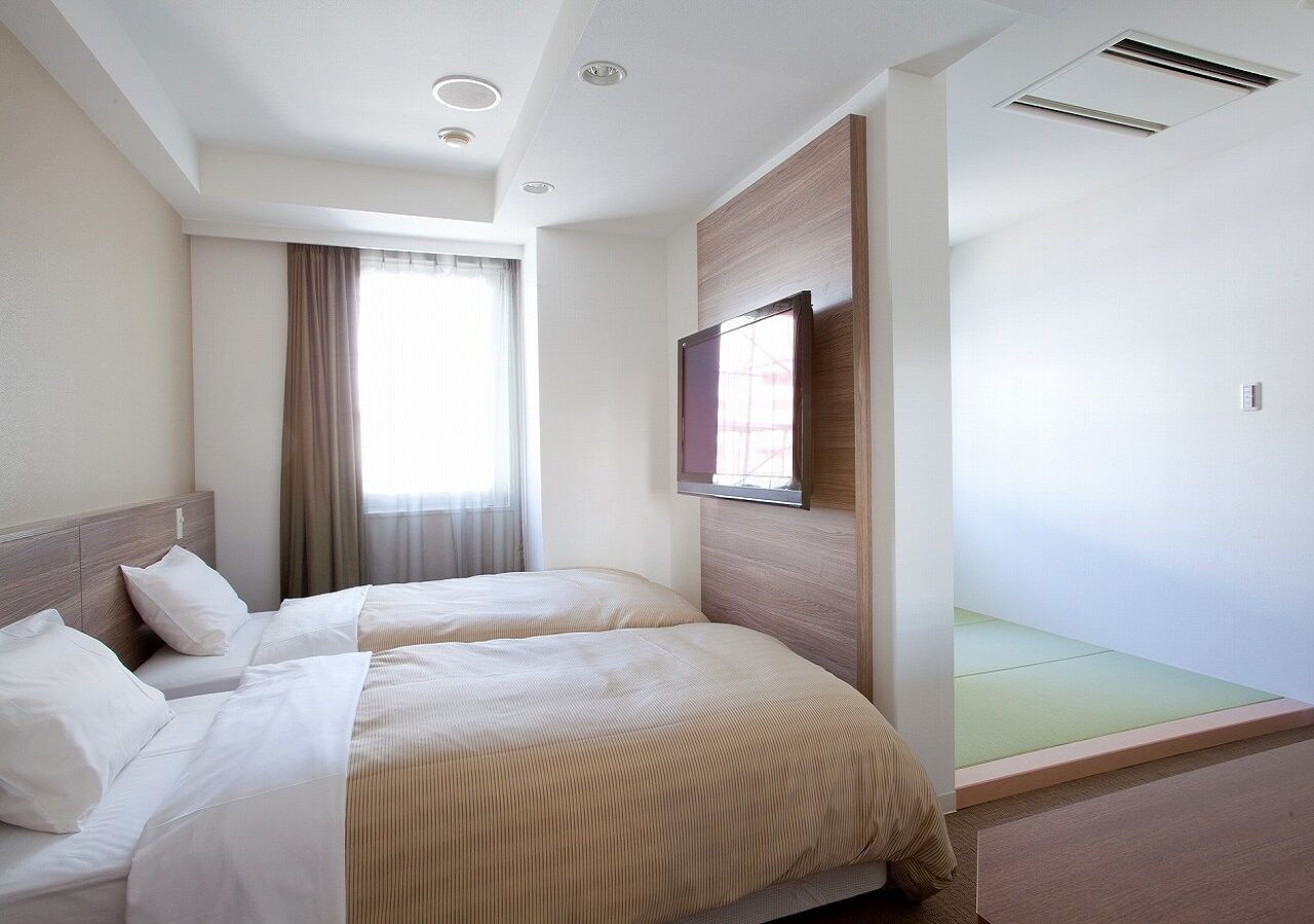 Habitación de hotel con dos camas, un ventanal grande con cortinas, un cabecero de madera y una pequeña zona de estar con un tapete verde.