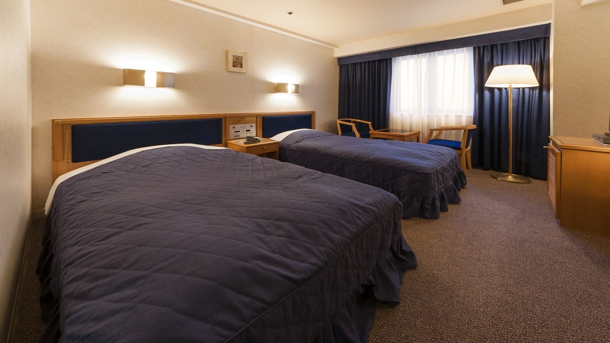 Habitación de hotel con dos camas, un escritorio, una silla, una lámpara y una ventana con cortinas.