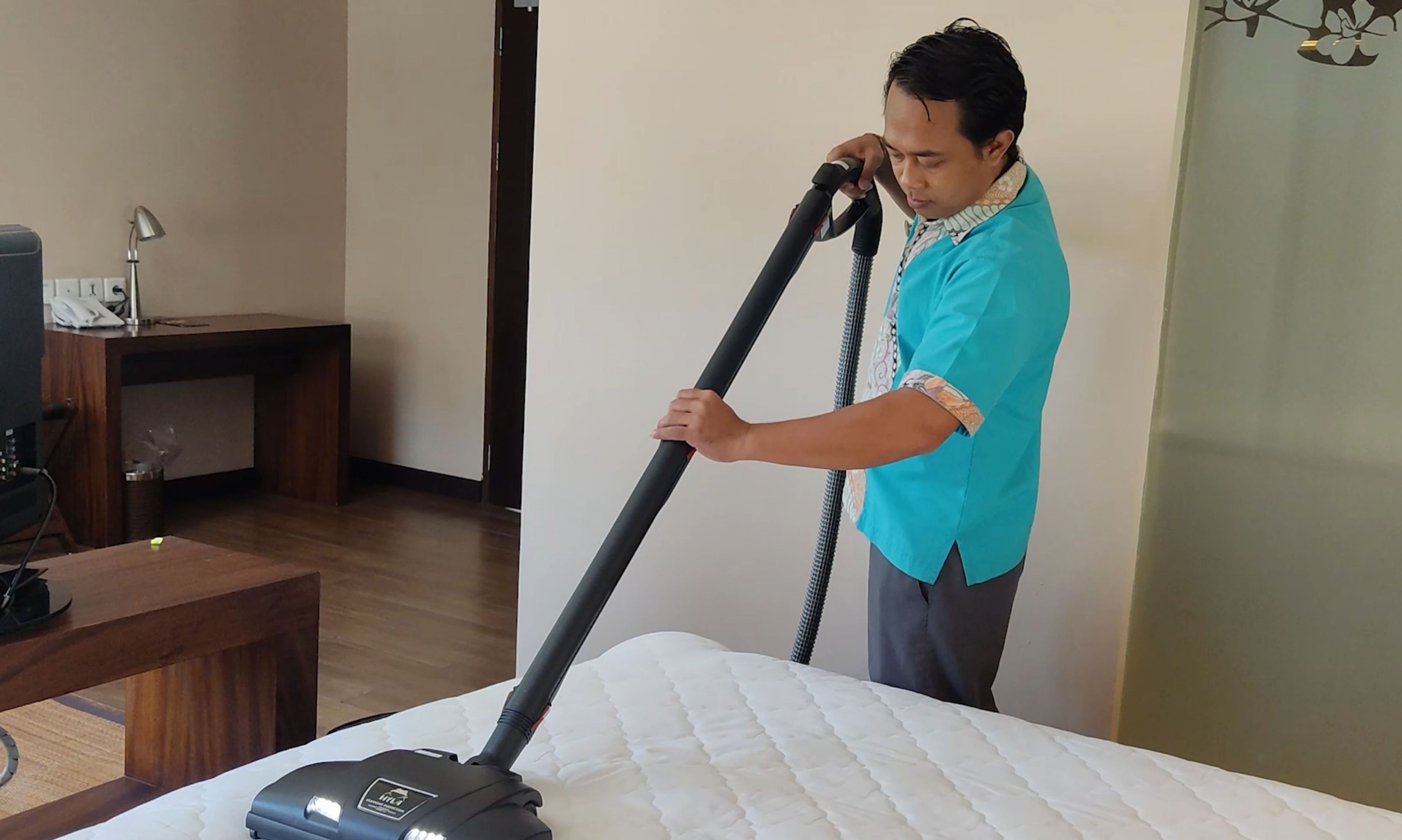 A person cleaning a bed with a vacuum cleaner in a room with a wooden floor, a desk, and a chair.