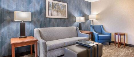 A hotel room with a grey sofa, a blue armchair, a small table, two lamps, and a framed picture on the wall.