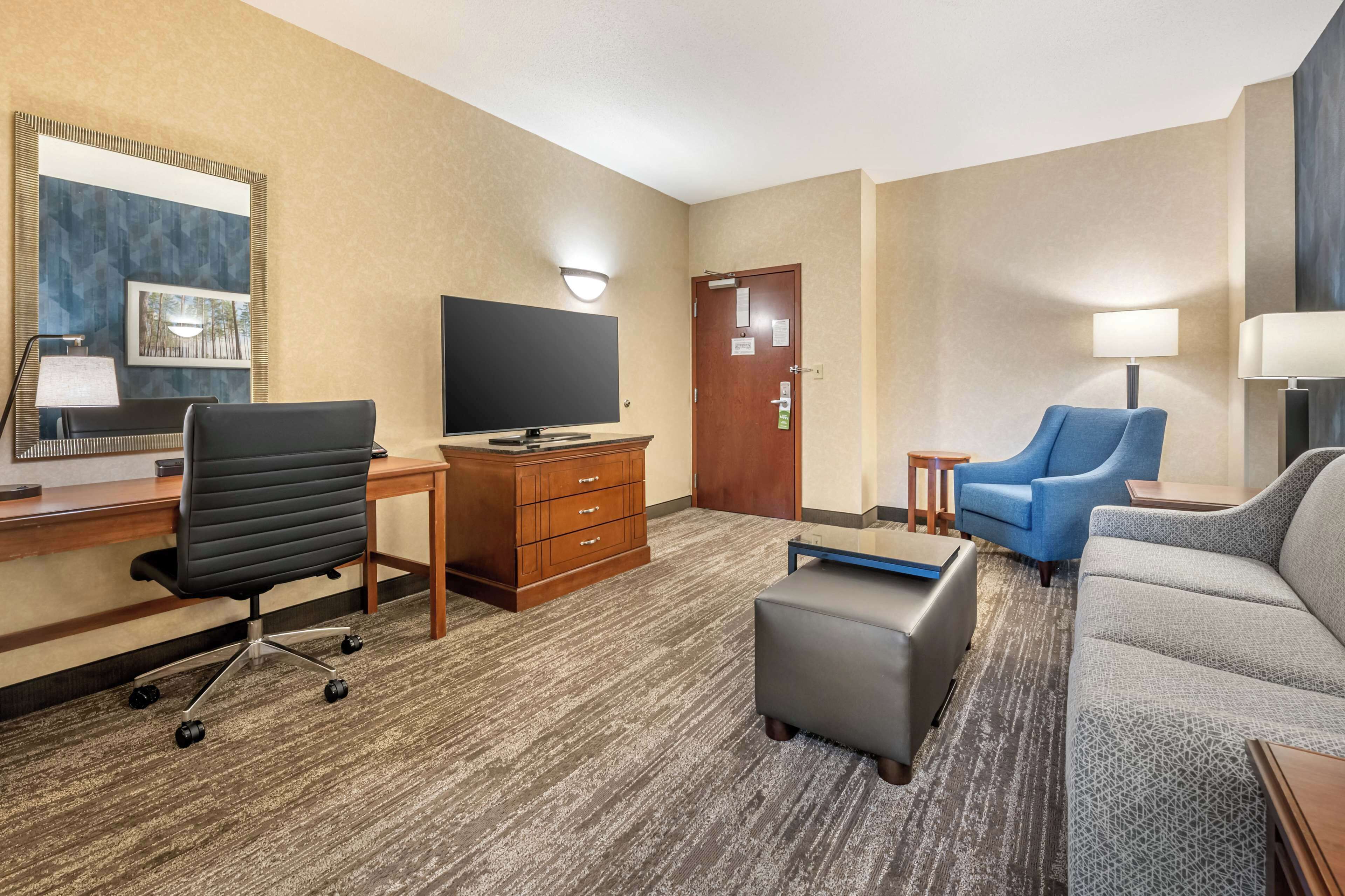 Una habitación de hotel con un escritorio, un televisor, un sillón azul, un sofá gris y un pequeño otoman.