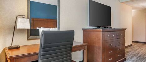 A hotel room with a wooden desk, a black office chair, a flat-screen TV on a wooden cabinet, and a large mirror on the wall.