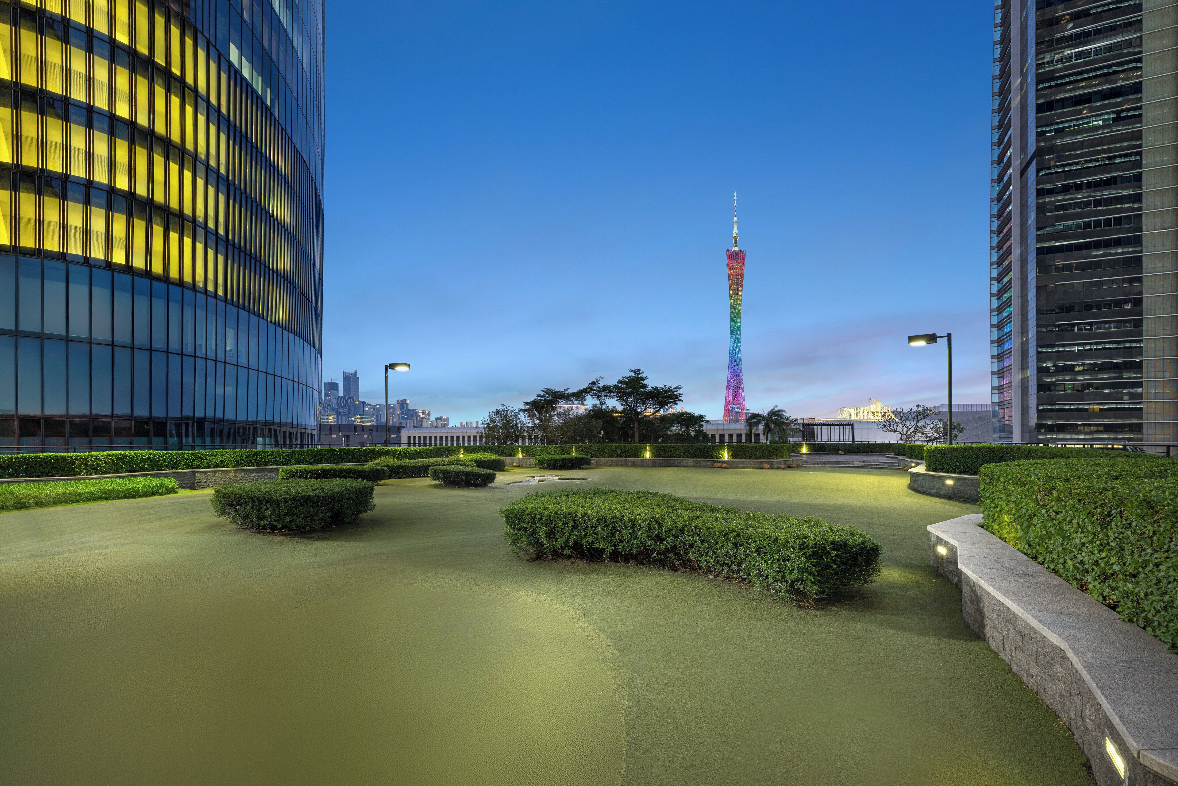 A modern urban plaza with a tall tower in the background, surrounded by well-maintained hedges and a water feature.