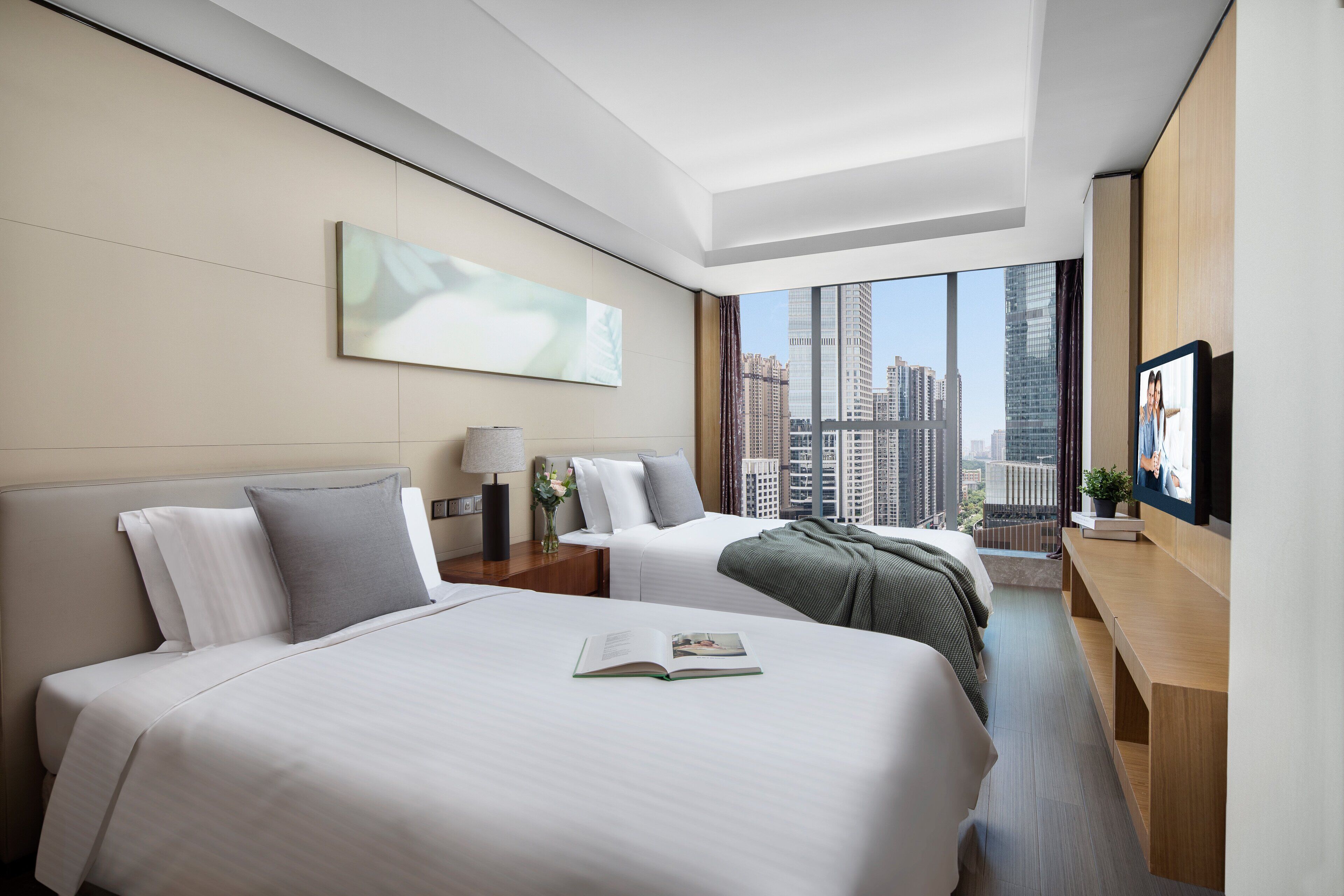 Habitación de hotel con dos camas, un escritorio, un televisor y un ventanal con vista a la ciudad.