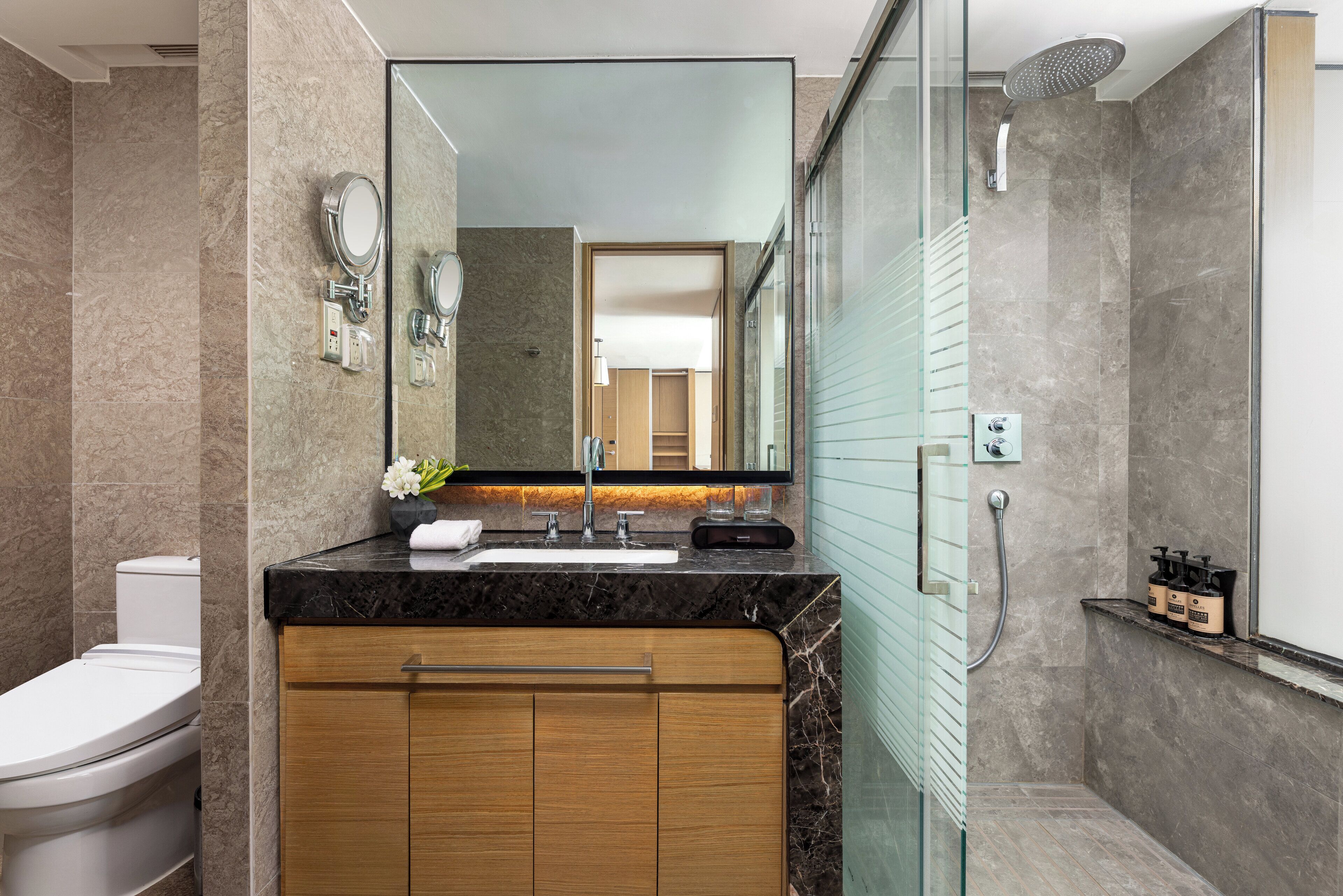 A modern bathroom with a marble sink, wooden cabinets, a glass shower enclosure, and a toilet.