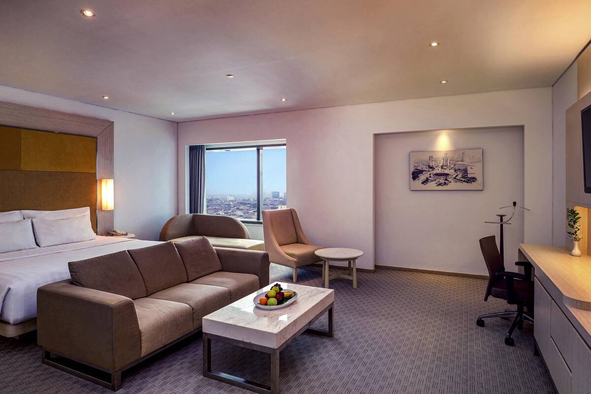 Hotelkamer met een bed, een slaapbank, een klein tafeltje met fruit, een stoel, een bureau en een groot raam met uitzicht op de stad.
