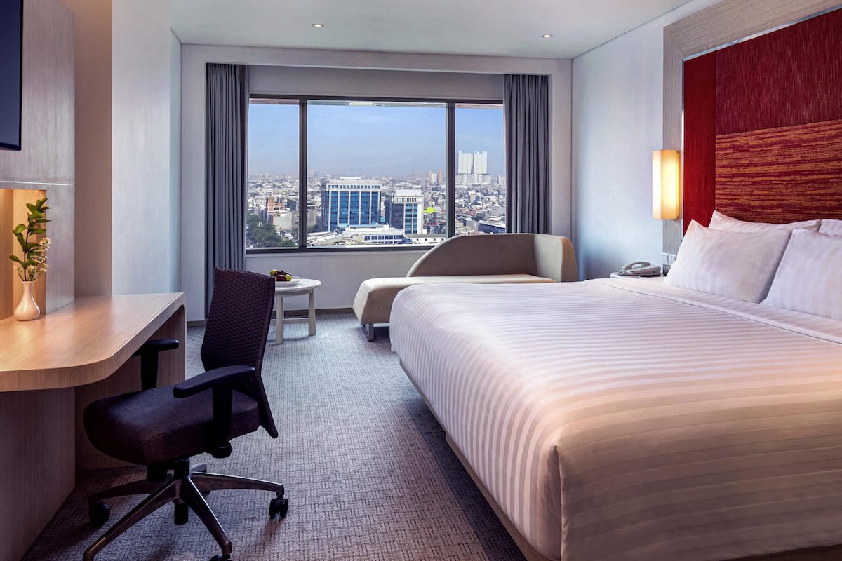 Une chambre d’hôtel avec un grand lit, un bureau, une chaise, une petite table et une vue sur le paysage urbain.