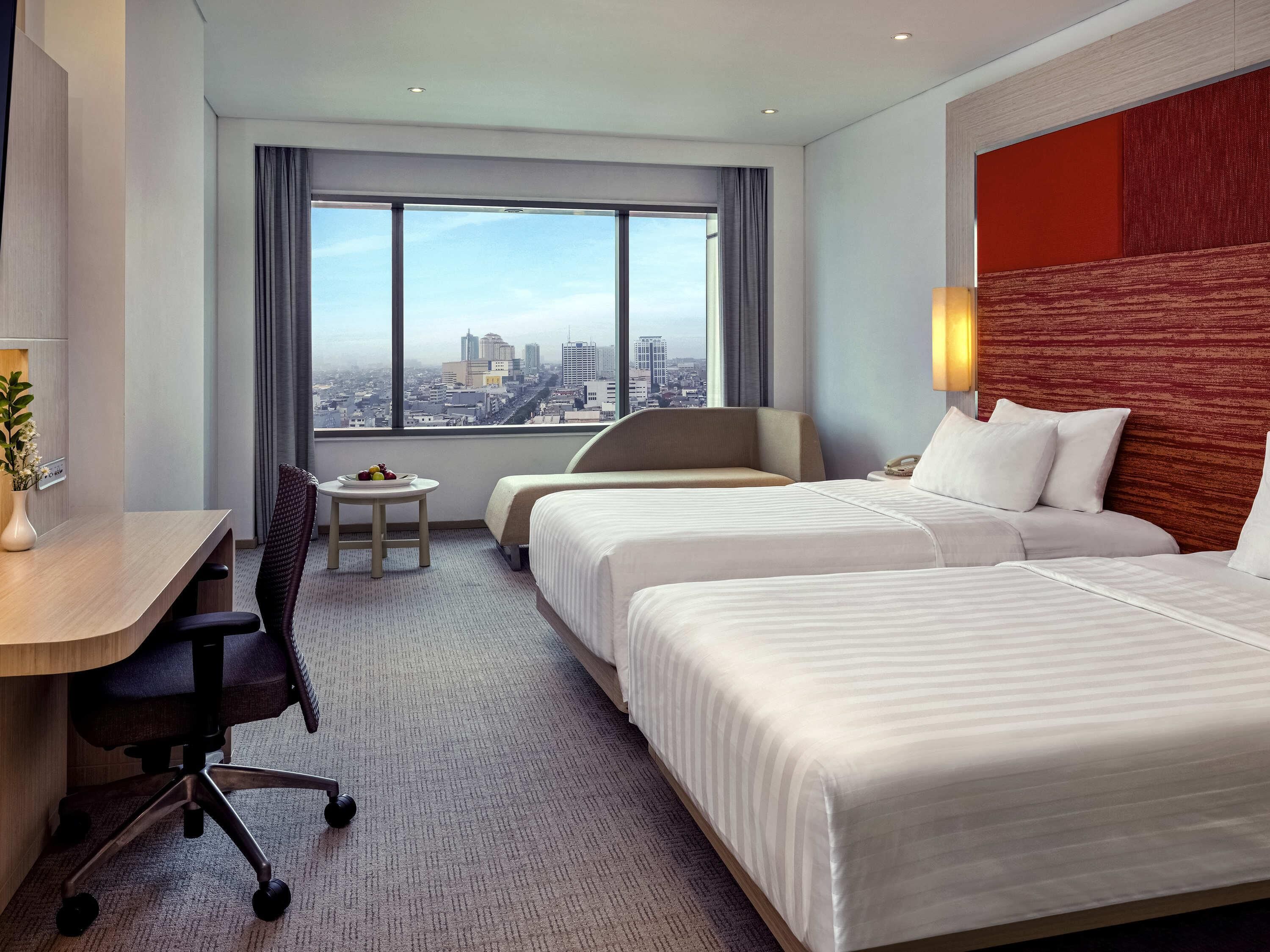 Een hotelkamer met twee bedden, een bureau, een stoel, een klein tafeltje met fruit, en uitzicht op de stad.
