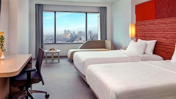 Habitación de hotel con vista a la ciudad, dos camas, un escritorio con silla y una pequeña mesa con fruta.