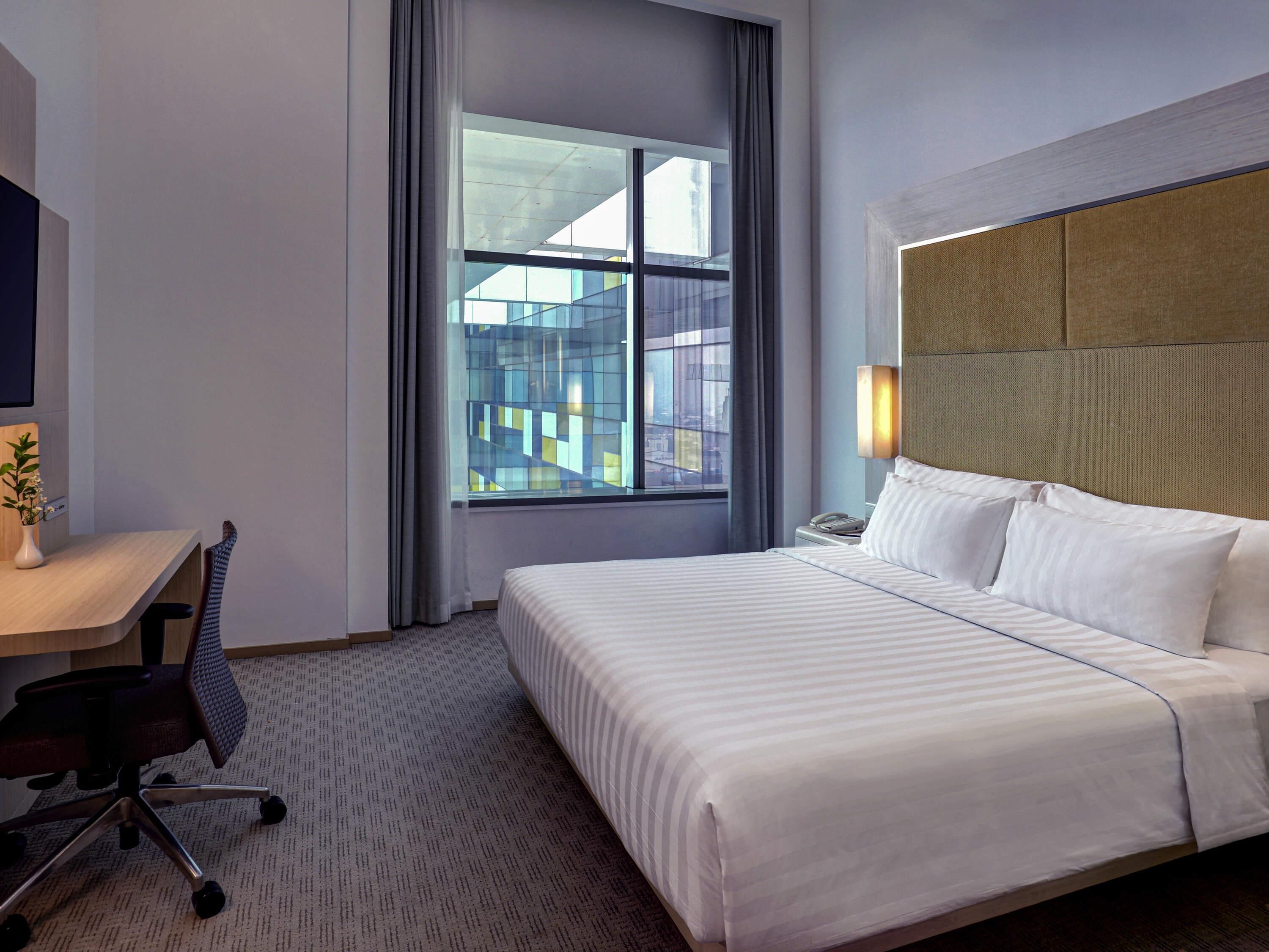 A hotel room with a large bed, a desk with a chair, a telephone, and a window with a view of buildings.