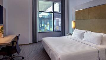 A hotel room with a large bed, a desk with a chair, a telephone, and a window with a view of buildings.