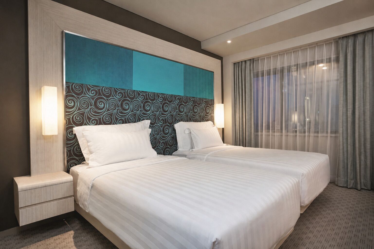 A hotel room with a large bed, two bedside tables, a patterned headboard, and a view through sheer curtains.