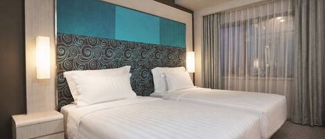 A hotel room with a large bed, two bedside tables, a patterned headboard, and a view through sheer curtains.