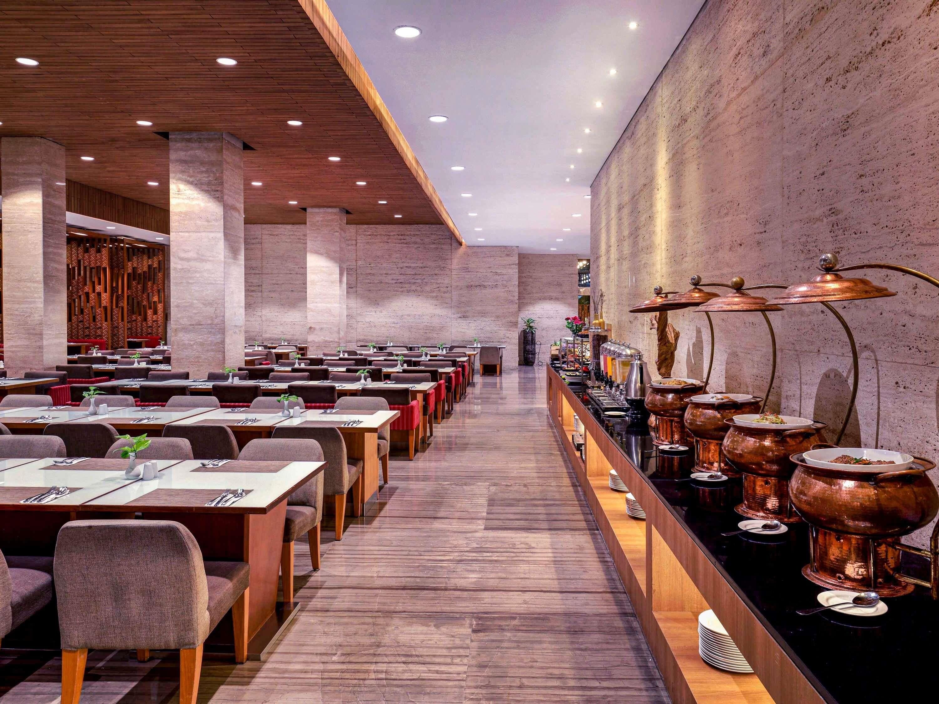 A spacious restaurant with multiple dining tables and chairs, a long counter with food service equipment, and a high ceiling with recessed lighting.