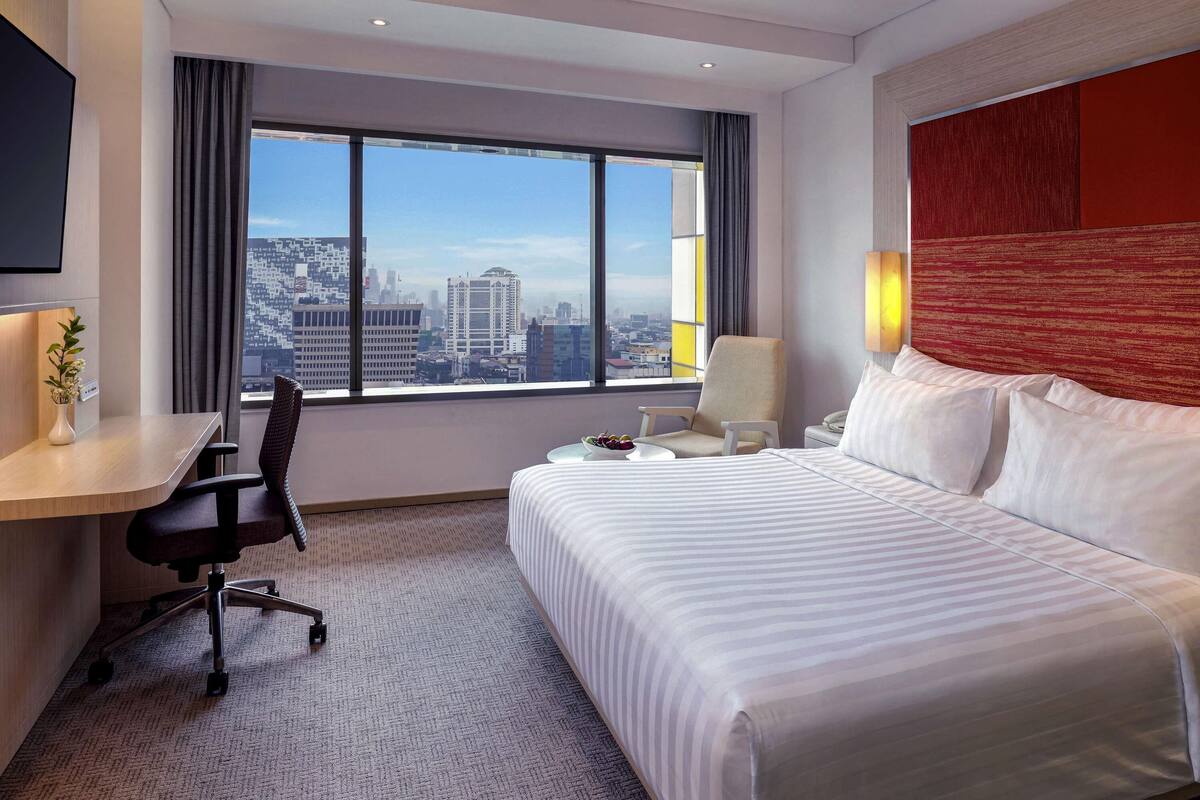 Een hotelkamer met een bed, bureau, stoel en een groot raam met uitzicht op de stad.