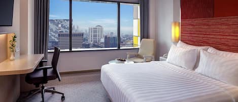 A hotel room with a bed, desk, chair, and a large window offering a city view.