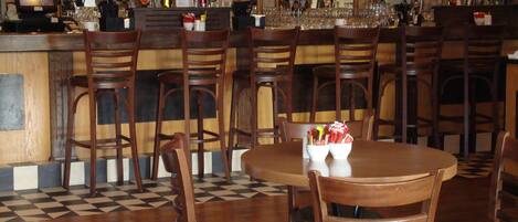 A bar area with wooden chairs and tables, a checkered floor, and a bar counter with various bottles and glasses.