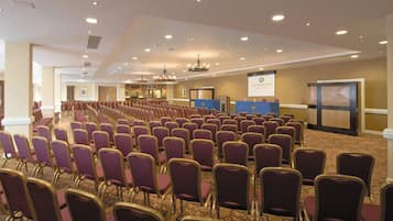 A conference room with rows of purple chairs, a stage with a podium, and a banner reading "ARD HOUSE HOTEL".