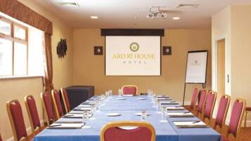 Un salón de conferencias en el Ard Ri House Hotel, con una larga mesa dispuesta para reuniones, sillas rojas, una pantalla de proyector y una pizarra.
