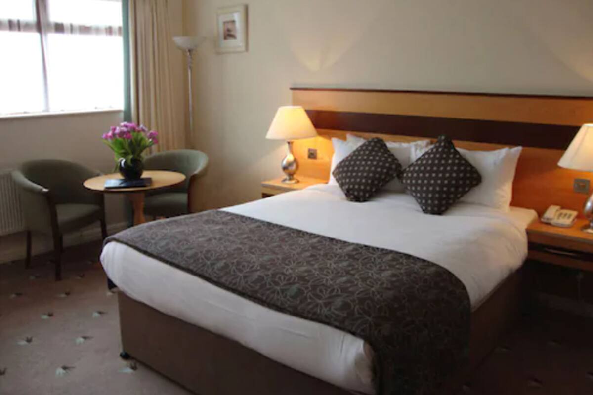 A hotel room with a large bed, two bedside tables with lamps, a chair, a small table with a vase of flowers, and a window with curtains.